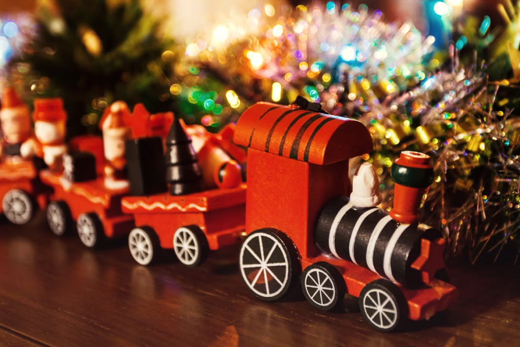 Wooden toy Christmas train with red cars and festive figures, displayed on a tabletop with colorful tinsel and blurred holiday lights in the background.