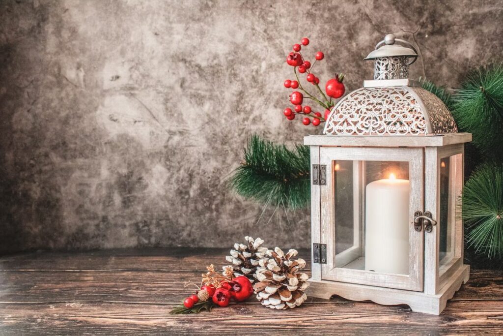 Rustic holiday lantern with a lit white candle, surrounded by evergreen branches, red berries, and pinecones on a wooden surface, creating a cozy DIY Christmas decoration scene.