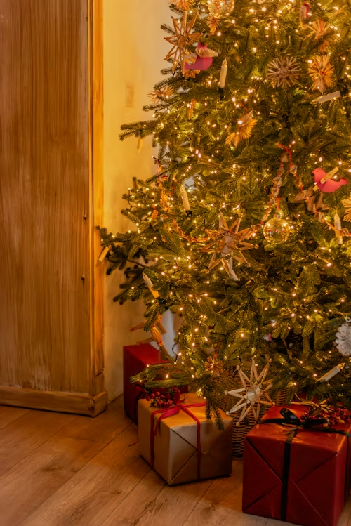 A warmly lit Christmas tree decorated with traditional Christmas tree colors, featuring natural straw ornaments, red accents, and warm white lights, with wrapped presents in red and gold beneath the tree on a wooden floor.