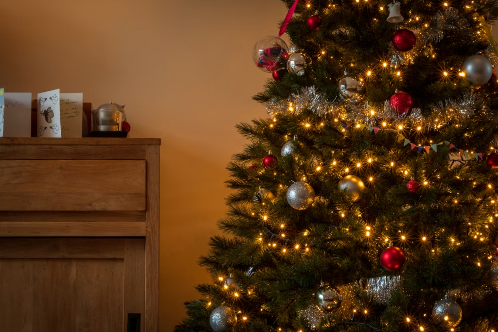 A Christmas tree decorated with traditional Christmas tree colors, featuring red and silver ornaments, warm white lights, and silver tinsel garland, set beside a wooden cabinet with holiday cards on top.