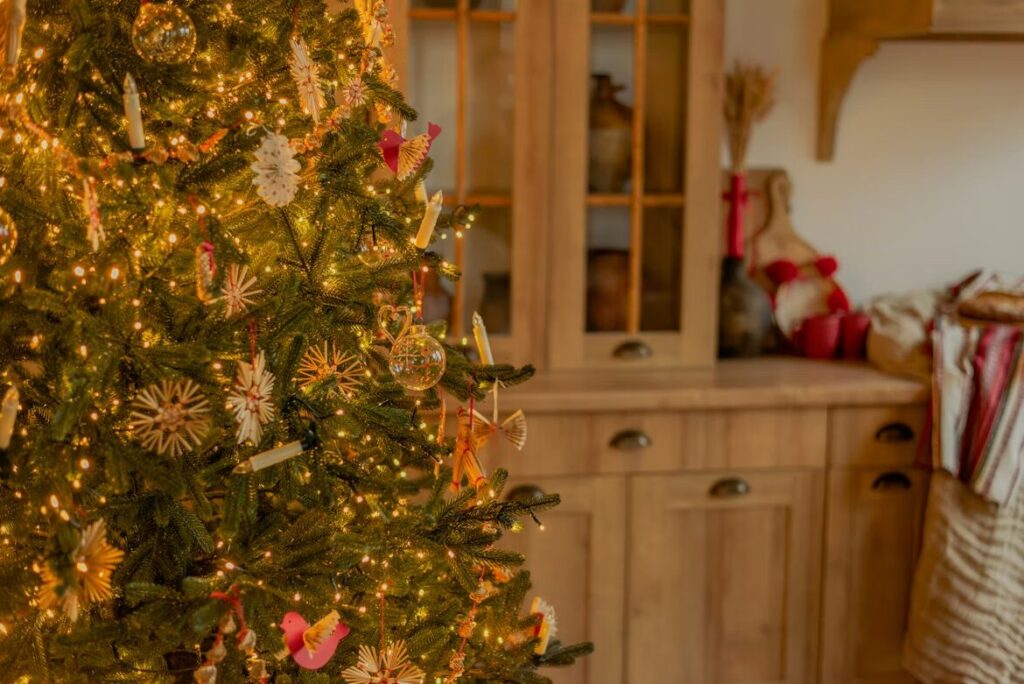 Close-up of a Christmas tree decorated with warm white lights and traditional Christmas tree colors, including natural straw ornaments, red accents, and handmade decorations, set in a cozy room with wooden cabinetry in the background.
