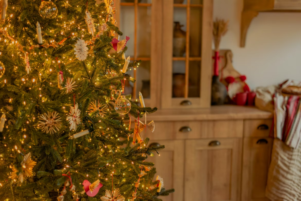 Close-up of a Christmas tree decorated with warm white lights and traditional Christmas tree colors, including natural straw ornaments, red accents, and handmade decorations, set in a cozy room with wooden cabinetry in the background.