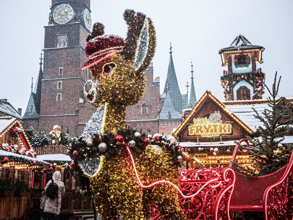A festive Christmas market scene with a large glittering reindeer decoration, red sleigh, and wooden stalls surrounded by snow, set against a historic European clock tower.