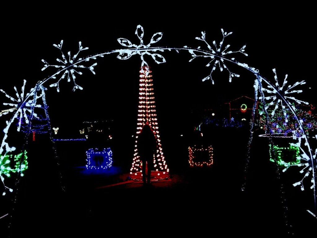 A person stands under a large illuminated arch decorated with white snowflake lights, facing a tall red Christmas tree display surrounded by colorful lighted shapes.