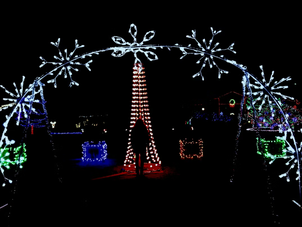 A person stands under a large illuminated arch decorated with white snowflake lights, facing a tall red Christmas tree display surrounded by colorful lighted shapes.