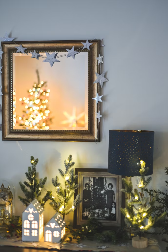 Holiday mantel decorated with mini evergreen trees wrapped in warm string lights, ceramic house decorations, framed photos, and a star garland around a mirror, creating a cozy DIY Christmas display.
