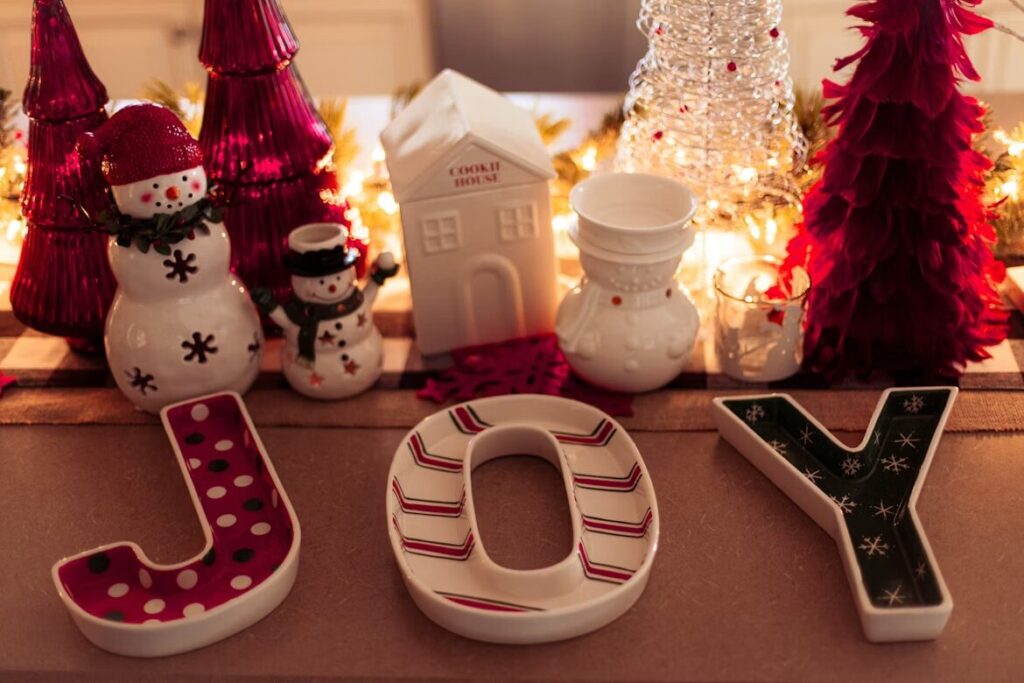Festive holiday tabletop display with ceramic letters spelling “JOY,” snowman figurines, decorative trees, a ceramic house, and warm string lights creating a cozy Christmas scene.
