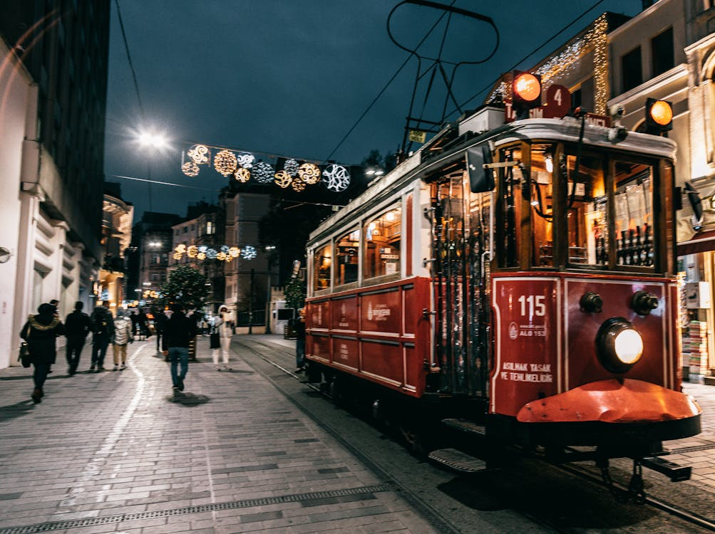 A vintage red tram decorated with lights travels down a festive, pedestrian street at night, with people walking under hanging holiday decorations.